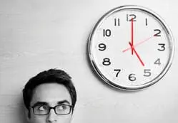 Foto de um homem olhando para um relógio ao seu lado esquerdo, o relógio marca 5 horas